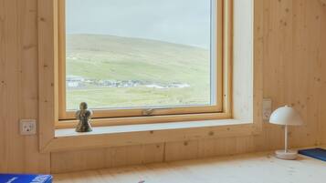 Una habitación con un escritorio de madera, una silla, un ventanal con vistas a un paisaje verde y una lámpara.