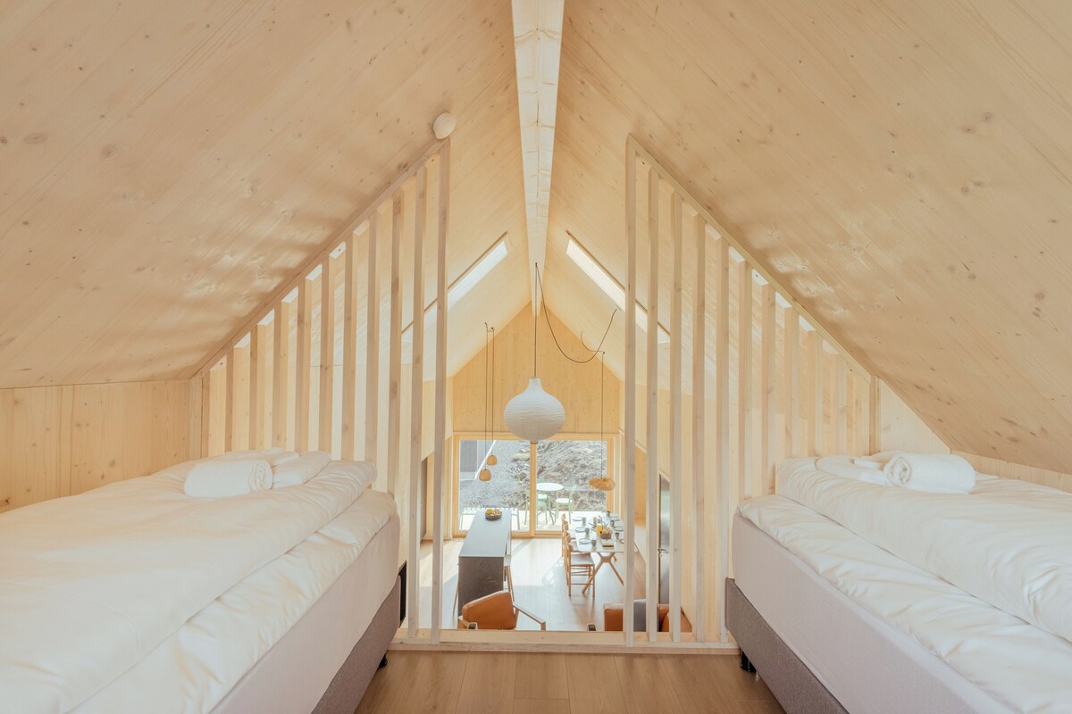 A high-ceilinged attic bedroom with two beds, a dining area, and large windows.