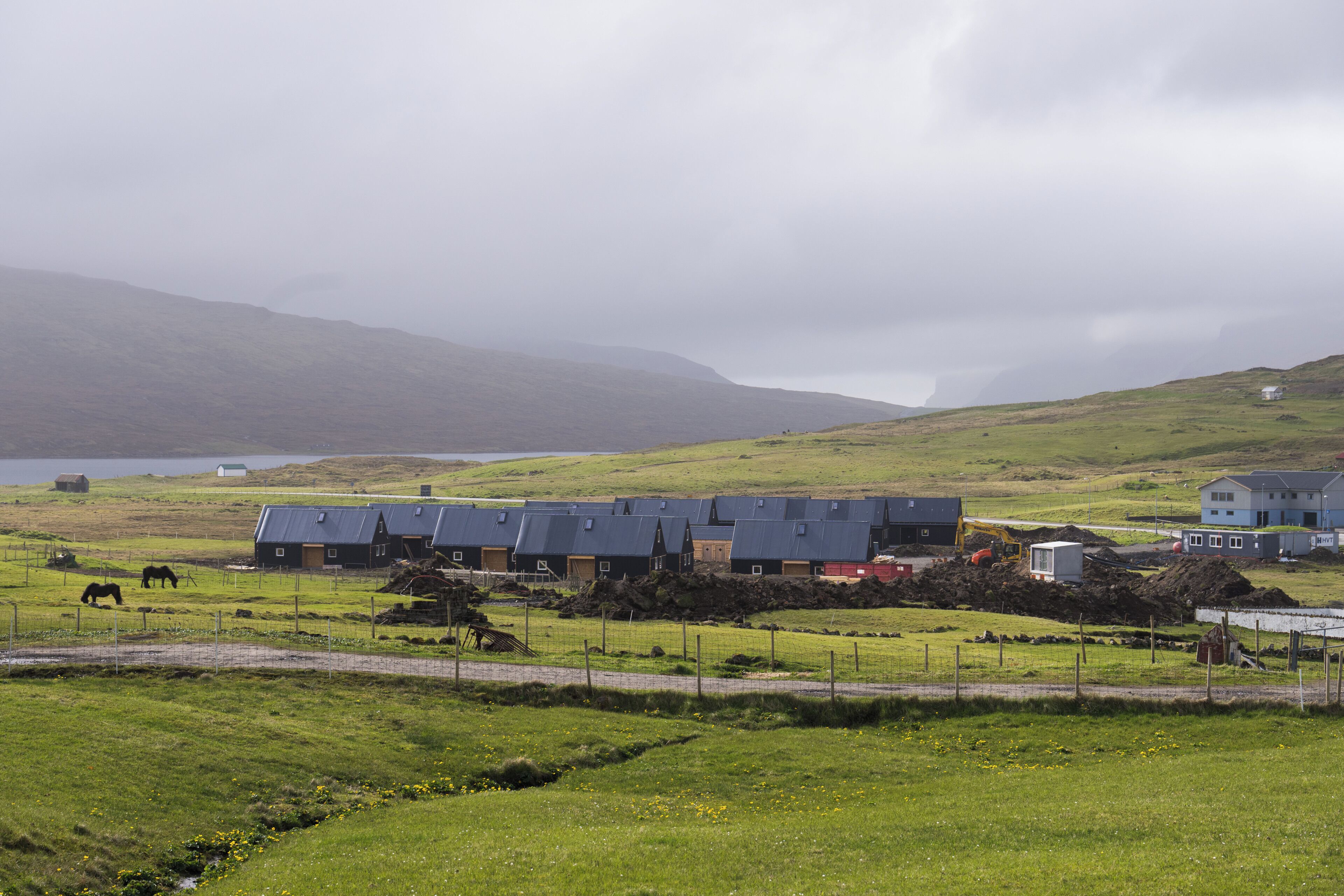 A rural landscape with a row of buildings, a fenced area with horses, and a body of water in the distance.