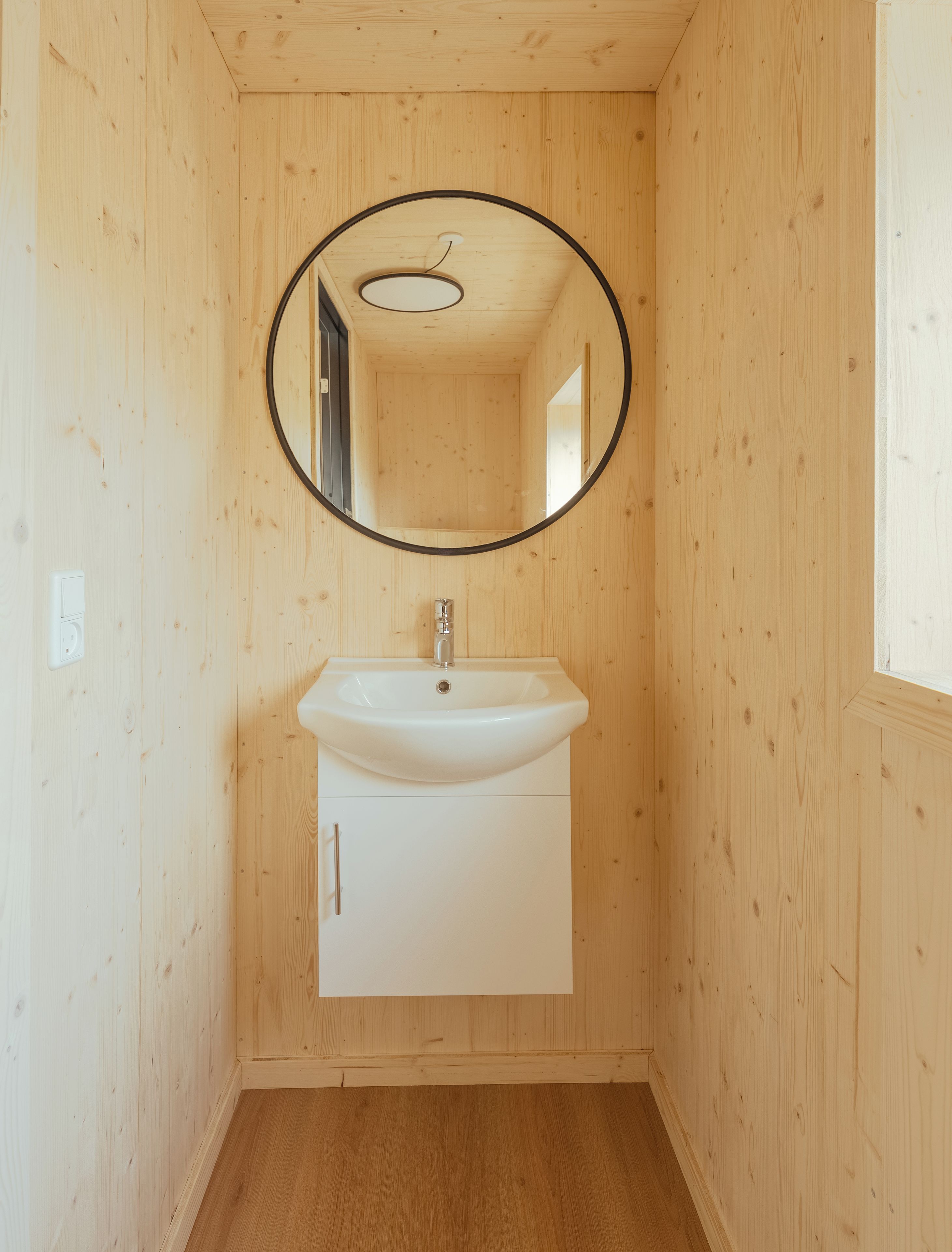 A wooden-paneled bathroom with a white sink, a round mirror, and a ceiling light fixture.