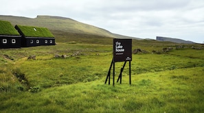 A sign for 'the Lakehouse' with a grassy field and a building with a grass roof in the background. - The Lakehouse (Miðvágur)
