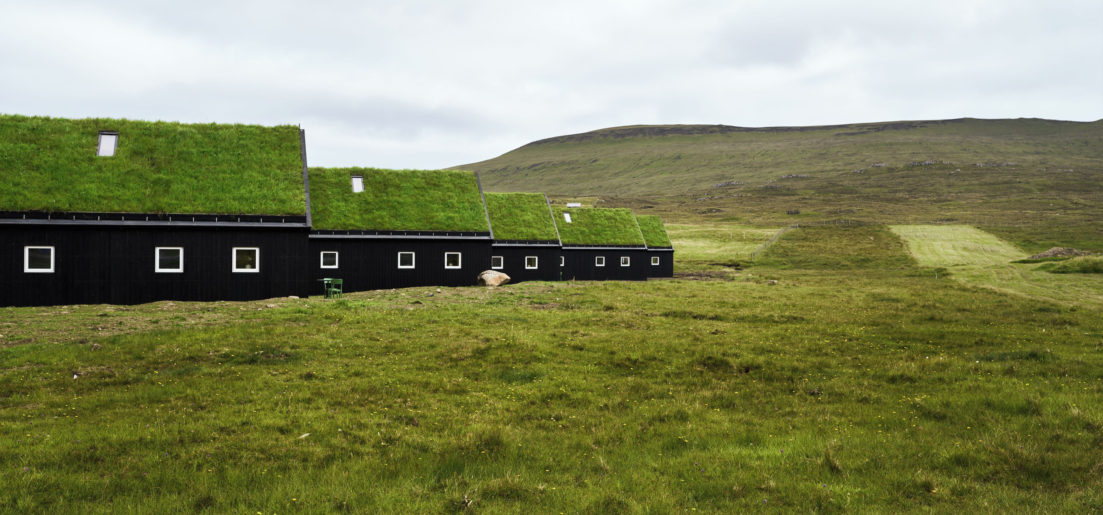A row of black buildings with grass roofs and small windows, set in a grassy field with a hill in the background.