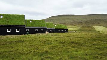 A row of black buildings with grass roofs and small windows, set in a grassy field with a hill in the background.