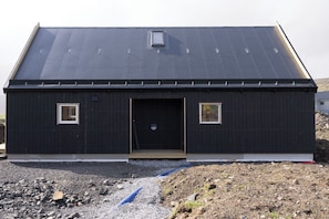 A single-story building with a dark exterior, a central door, and two small windows on either side. - The Lakehouse (Miðvágur)