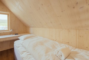 A room with a wooden sloped ceiling, a bed with white bedding, a desk with a lamp, and a window with a view of a building and landscape. - The Lakehouse (Miðvágur)