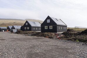 A row of black buildings with sloped roofs, gravel ground, and a cloudy sky. - The Lakehouse (Miðvágur)
