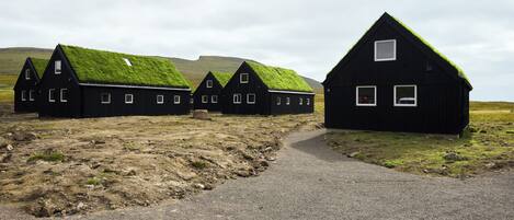 A row of black buildings with grass roofs, a gravel path, and a hilly landscape.