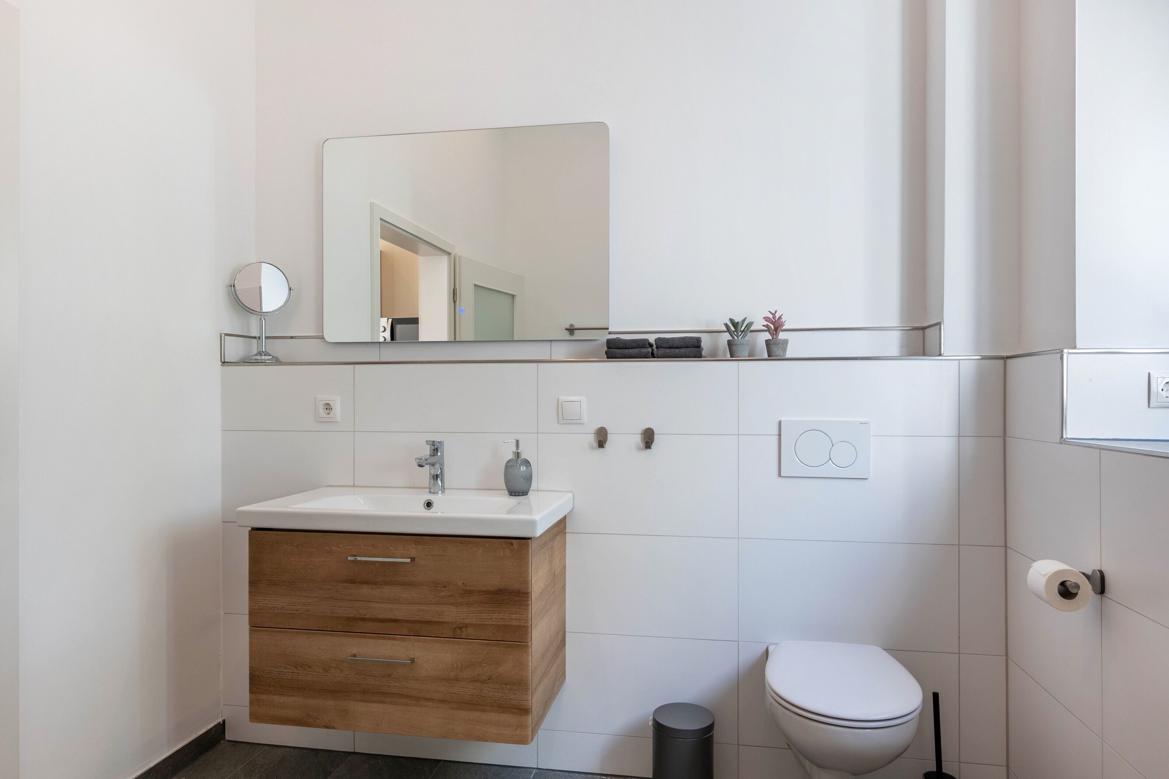 A modern bathroom with a wooden vanity, a mirror, a toilet, and a shelf with toiletries.