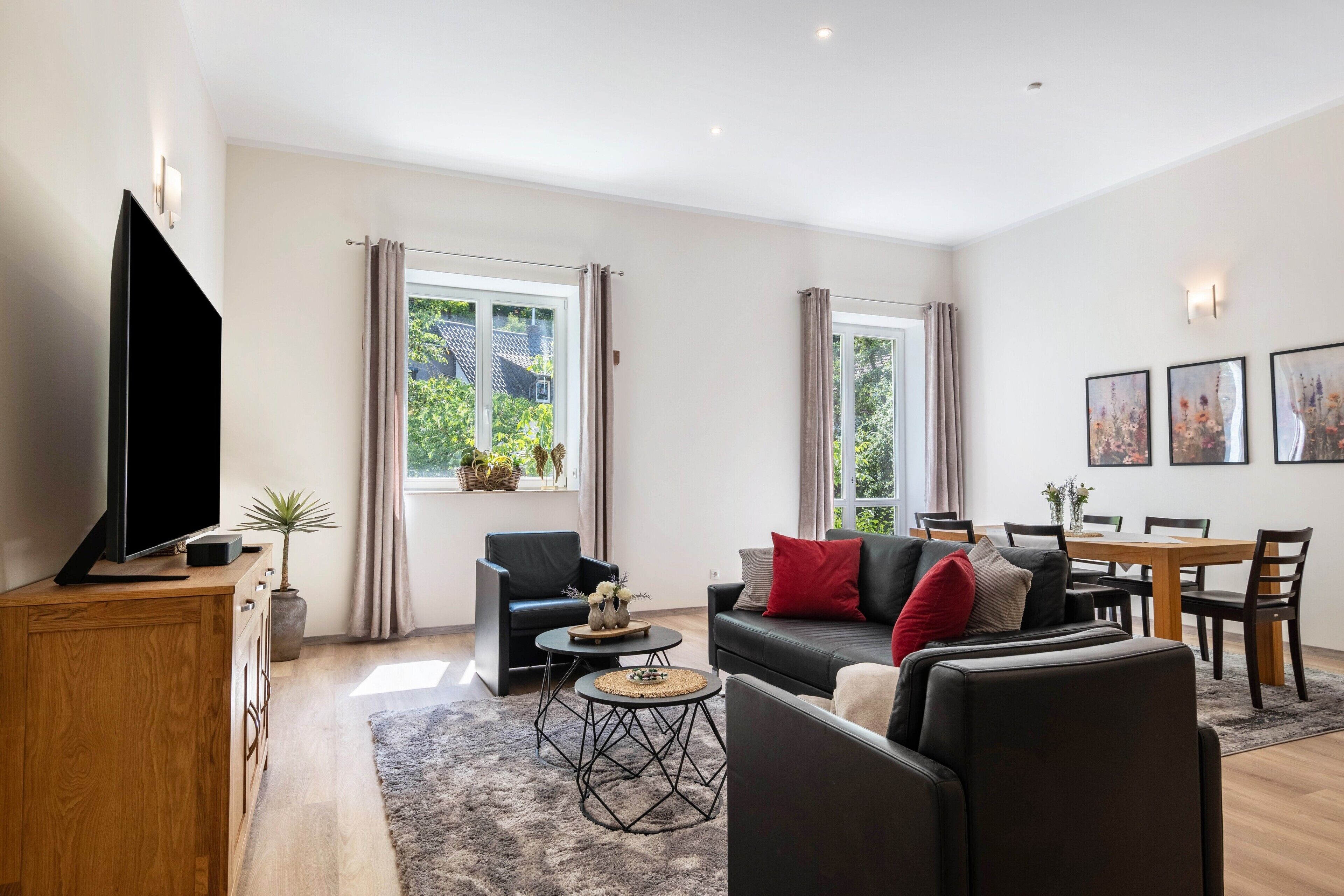 Ein modernes Wohnzimmer mit Flachbildfernseher, einer Couch, einem Esstisch und einem großen Fenster mit Vorhängen.