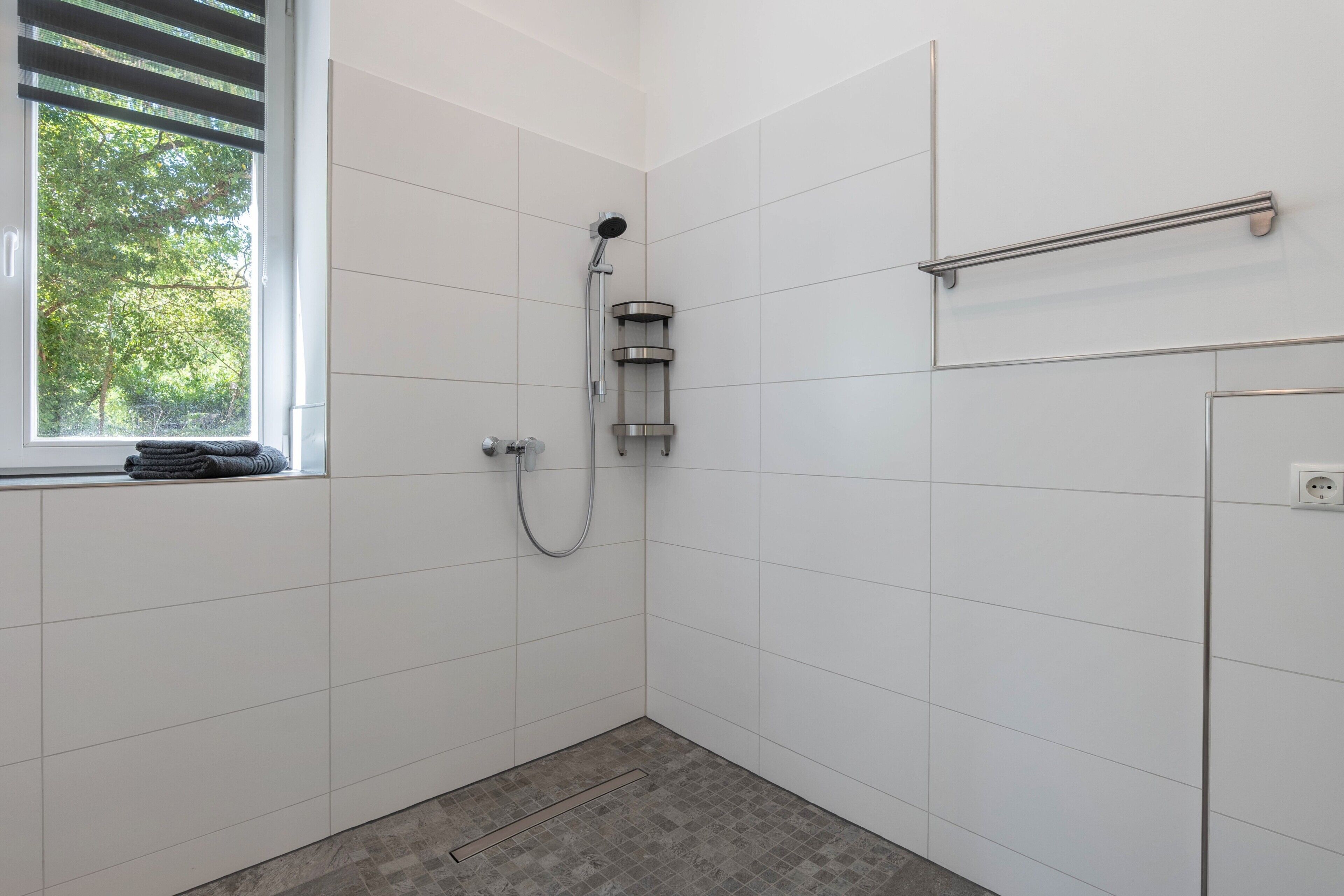 A small, modern bathroom with white tiles, a shower, a window with blinds, and a towel rack.