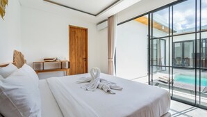 A hotel room with a large bed, a wooden door, a small table with a lamp, and a view of a swimming pool through large windows. - VILLA TRATASAN CANGGU (Badung)