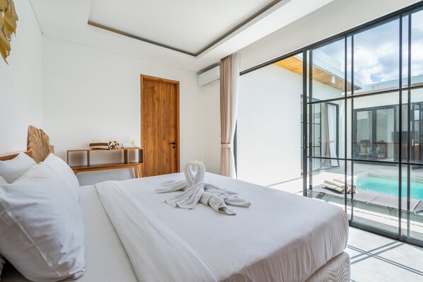 A hotel room with a large bed, a wooden door, a small table with a lamp, and a view of a swimming pool through large windows. - VILLA TRATASAN CANGGU (Badung)