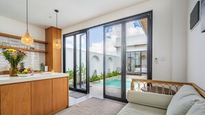 A modern kitchen with a central island, a dining area with a glass table, and a large sliding glass door leading to a pool area. - VILLA TRATASAN CANGGU (Badung)