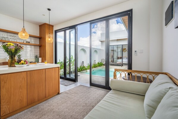 Una cocina moderna con una isla central, un comedor con mesa de vidrio y un amplio ventanal corredera que da a una zona de piscina.