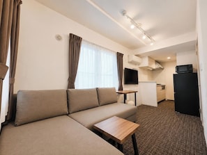 A compact living space with a sofa, a small table, a TV, a kitchenette, and an air conditioning unit.