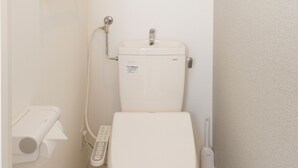 Een kleine, moderne badkamer met een toilet, een hygiëneschuimer, een toiletrolhouder en een plank met toiletpapier.