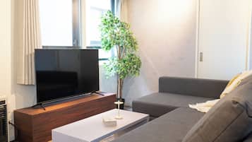 A modern living room with a flat-screen TV on a wooden stand, a grey sofa, a white coffee table with a book, and a potted plant.