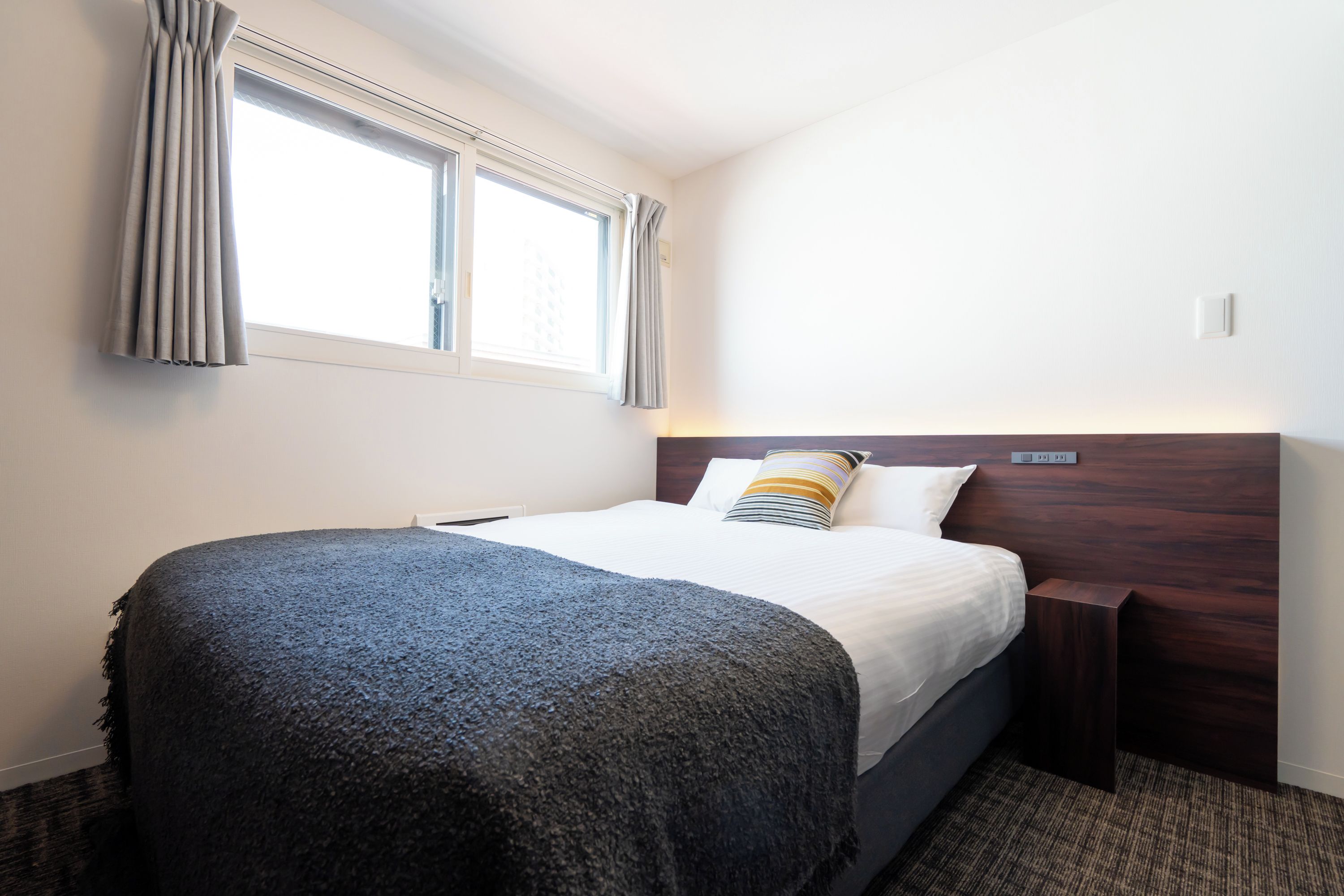 Una habitación de hotel con cama, mesita de noche, ventana con cortinas y un interruptor de luz en la pared.