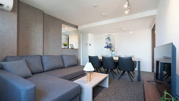 Una sala moderna con un sofá gris, una mesa de centro y un televisor. Se observa un área de comedor con una mesa y sillas al fondo.