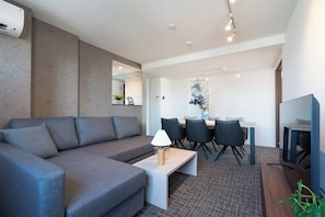 A modern living room with a grey sofa, a coffee table, and a TV. There is a dining area with a table and chairs in the background. - Hotel Luxe HIRAGISHI (Sapporo-shi)