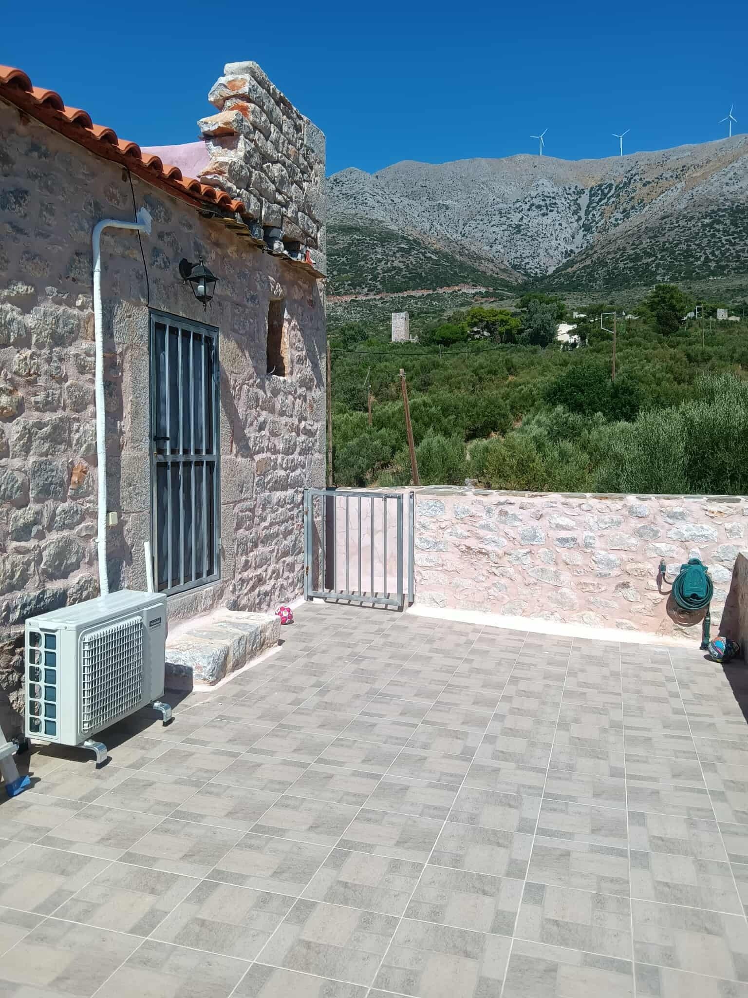 Un edificio de piedra con un patio empedrado, una ventana con rejas y vistas a aerogeneradores a lo lejos.