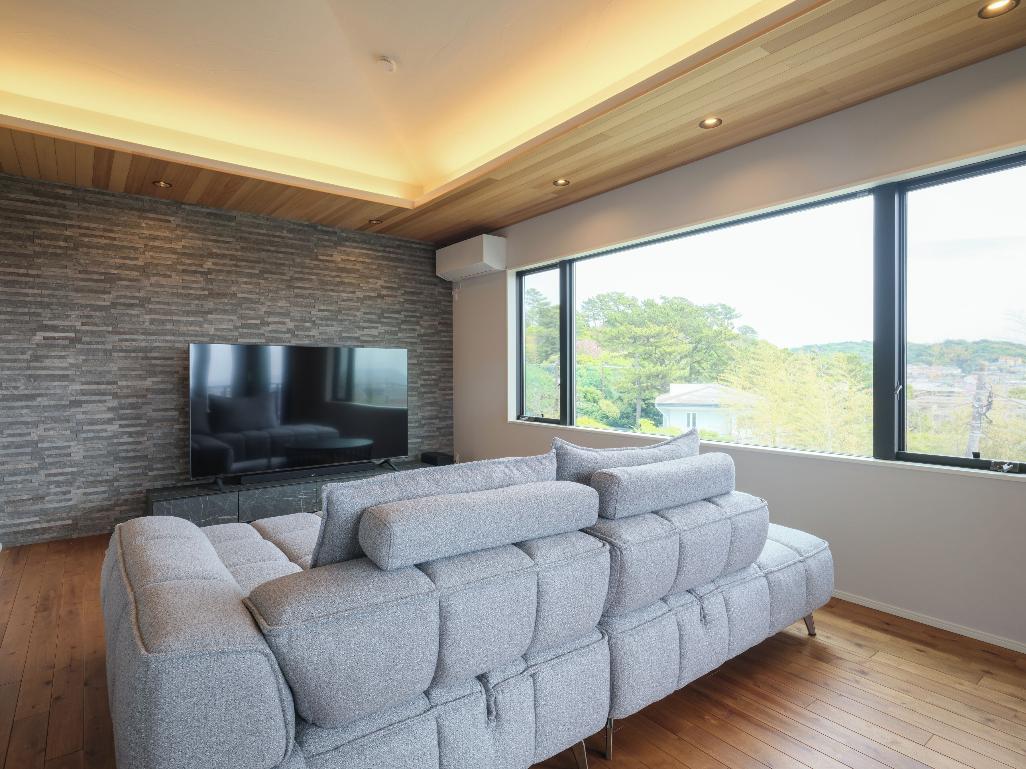 A modern living room with a large flat-screen TV, a grey sofa, and a wooden ceiling.