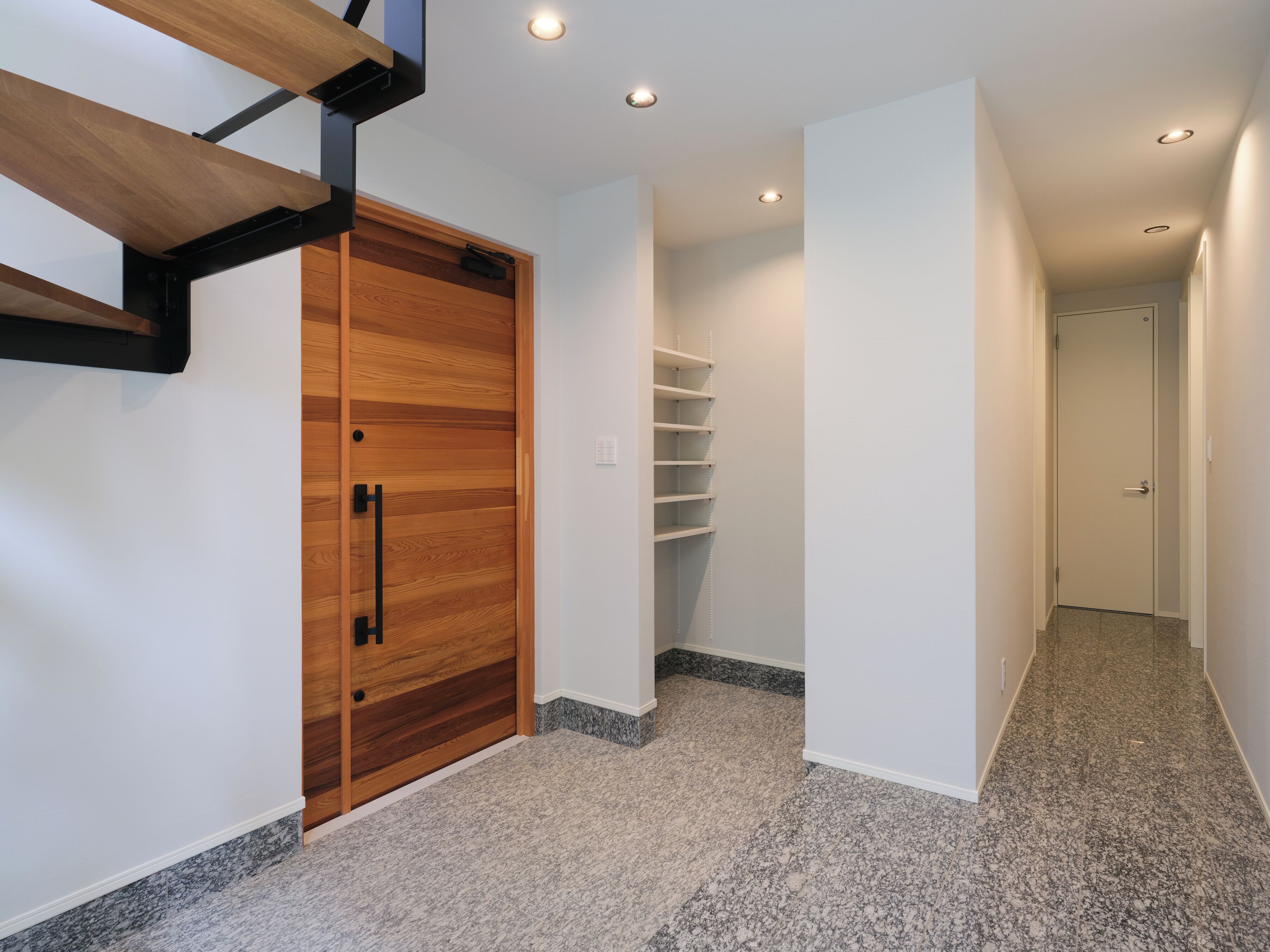 Un couloir avec une porte en bois, un mur blanc, une étagère et un sol recouvert d’une moquette.