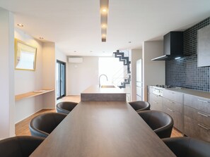 A modern kitchen with a central island, black chairs, a staircase, and a view to the living area. - KAMAKURA VILLA SAUNA KOSHIGOE 5 (Koshigoe, Kamakura-shi)