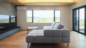 Ein modernes Wohnzimmer mit einem Ecksofa, einem runden Couchtisch und großen Fenstern mit Blick ins Freie.