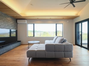 A modern living room with a sectional sofa, a round coffee table, and large windows offering a view of the outdoors. - KAMAKURA VILLA SAUNA KOSHIGOE 5 (Koshigoe, Kamakura-shi)