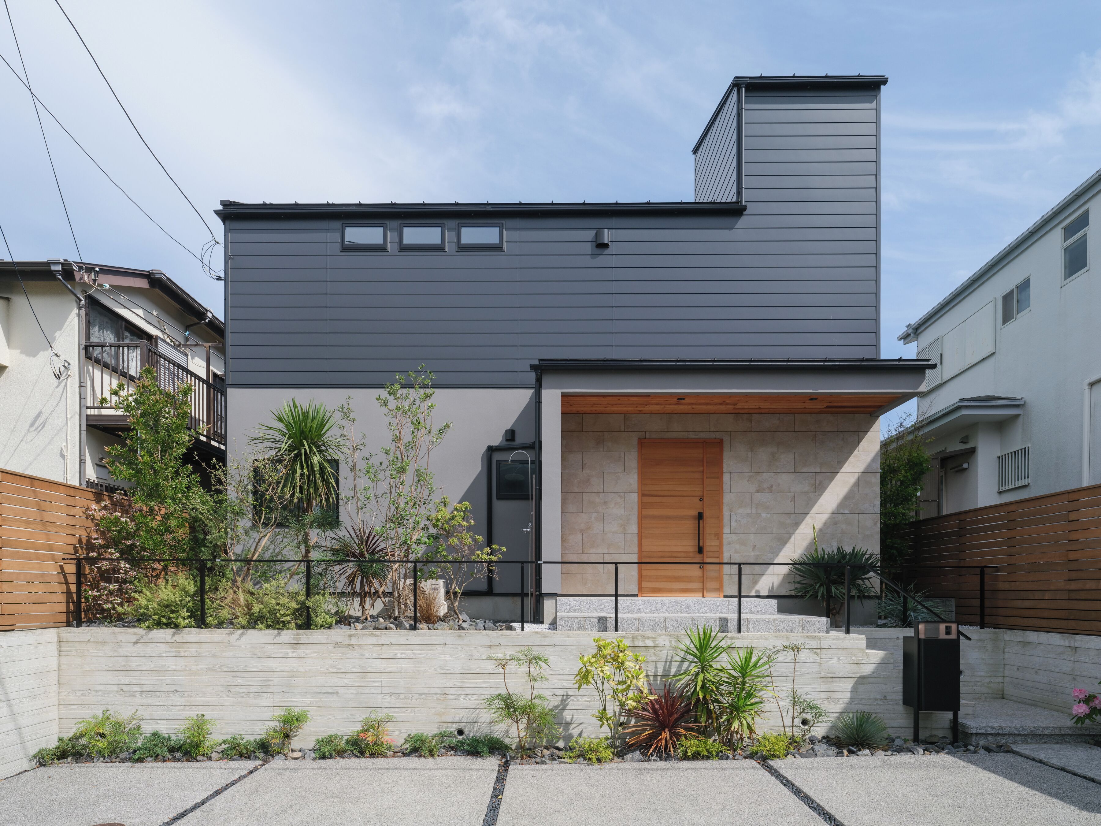 A modern two-story house with a grey exterior, a wooden front door, and a small garden with various plants.