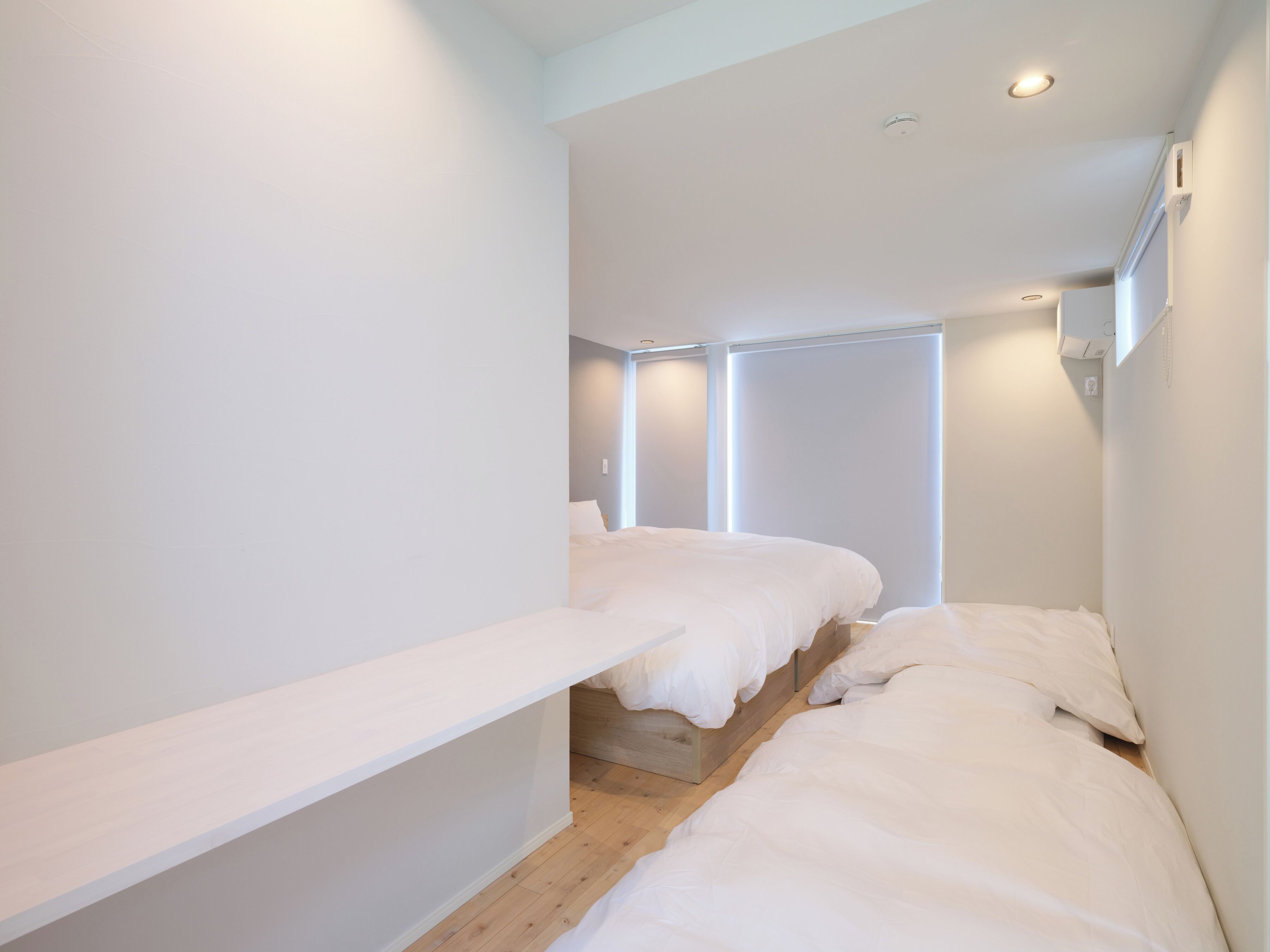 A narrow hallway with two beds, wooden flooring, white walls, and recessed ceiling lights.