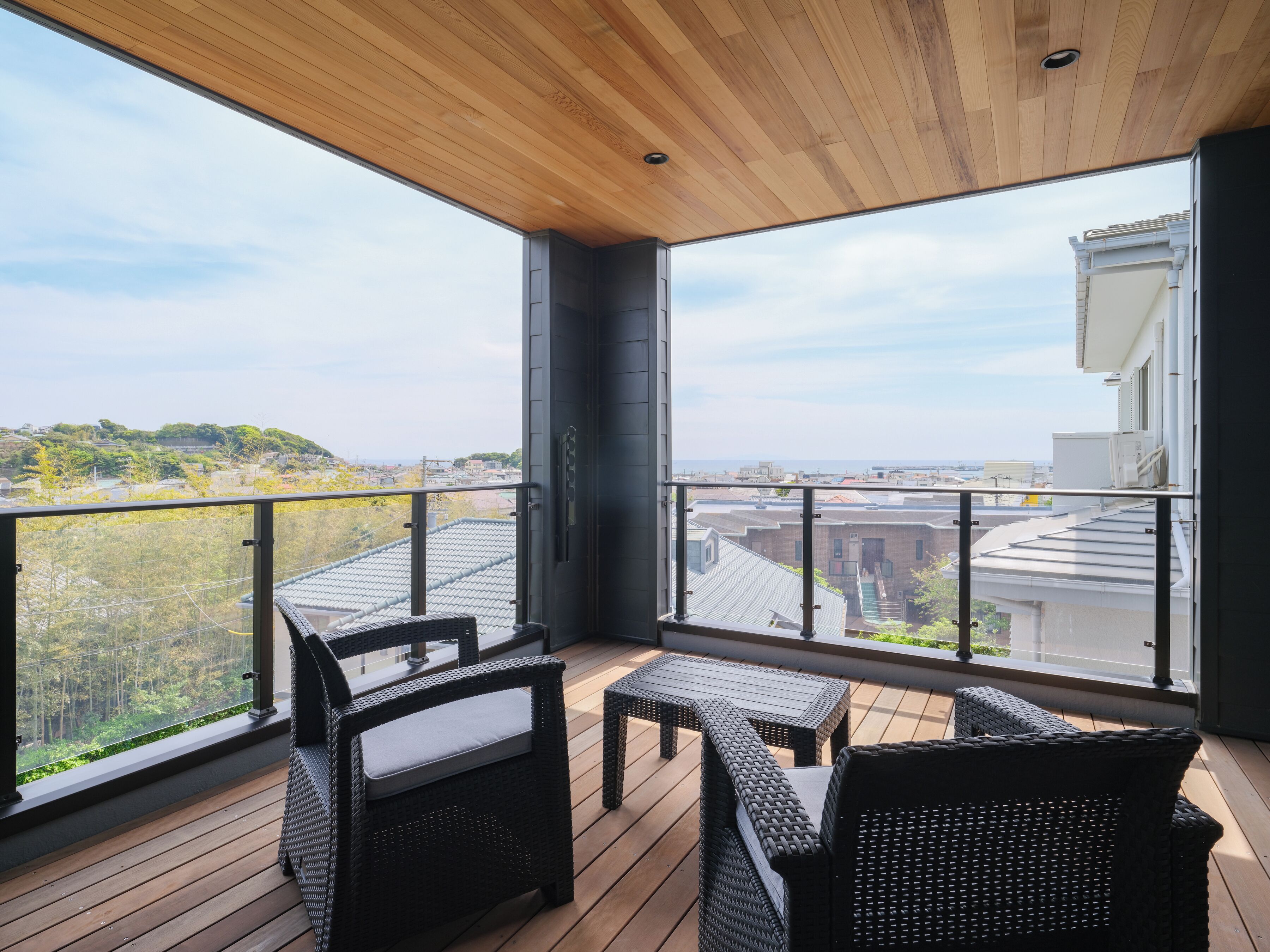 A balcony with wicker furniture, a wooden ceiling, and a view of buildings and greenery.