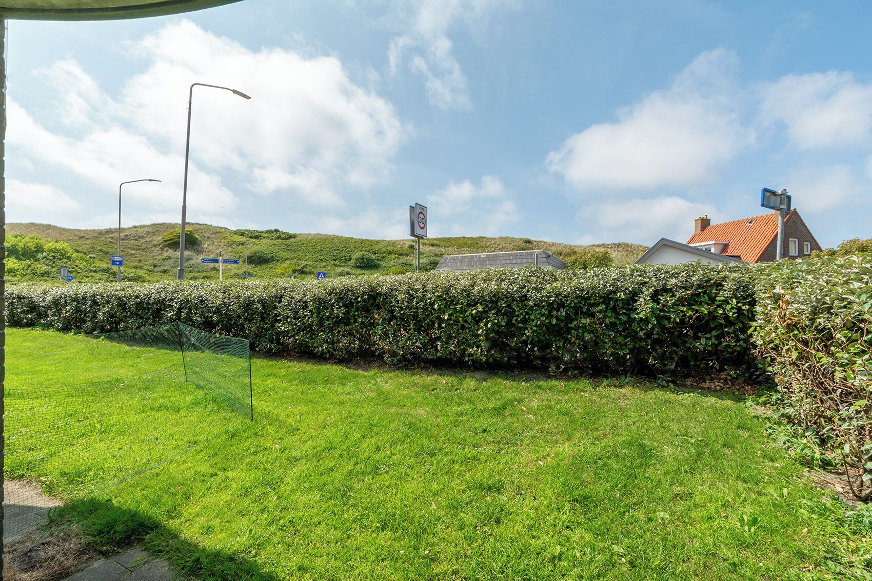 A grassy area with a hedge, a house with a red roof, and a no parking sign.