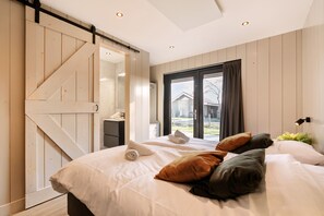 A bedroom with a large bed, a sliding barn door, a window with a view, and a bedside table with a lamp. - Zee Vacation Home – Luxury, Peace, and Space on the Coast (Julianadorp)