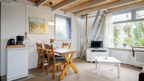 A cozy room with a wooden dining table, chairs, a small white cabinet, a television, and a window with a view of greenery. - Cozy vacation home for two, close to the beach, the woods, and tulip fields (Callantsoog)