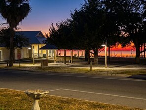 Een gebouw met een overdekte veranda, een straat op de voorgrond en een zonsondergang op de achtergrond.
