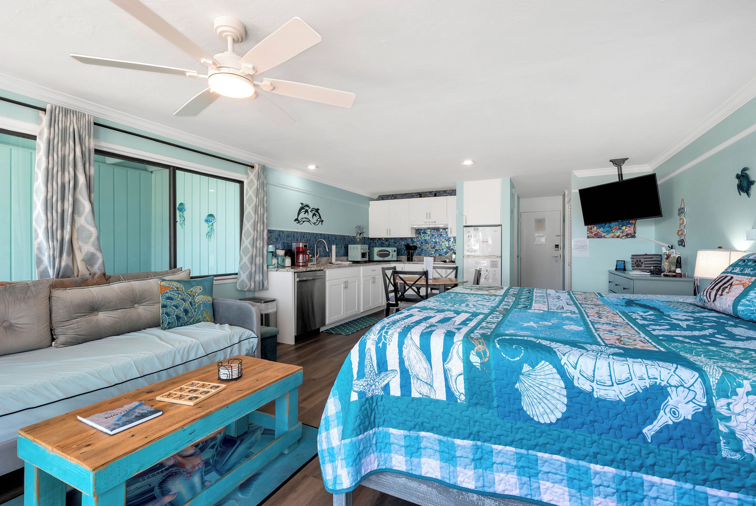 A compact living space with a kitchenette, a ceiling fan, a bed with a blue and white patterned bedspread, and a sofa with a turtle pillow.