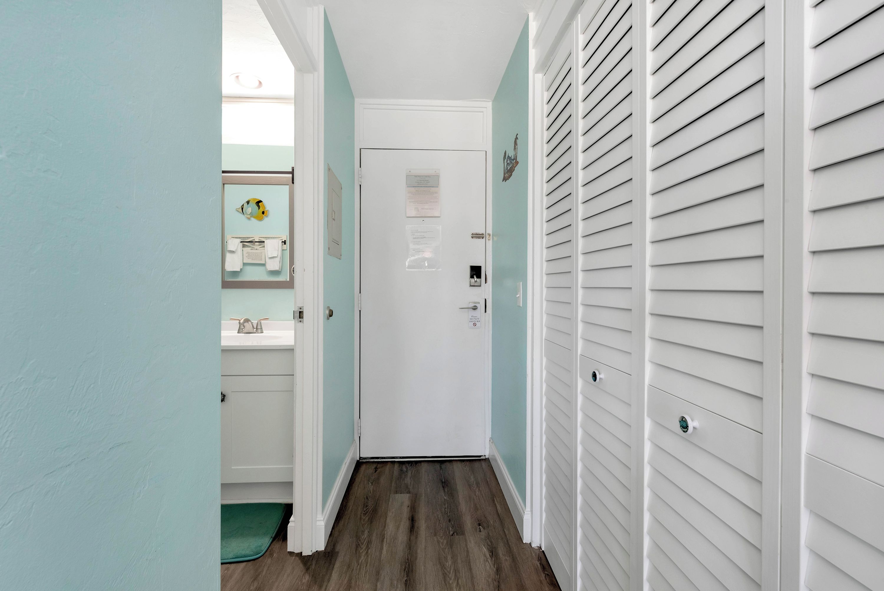 A narrow hallway with a door at the end, a small bathroom with a sink and towel rack, and a green mat at the entrance.
