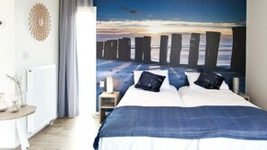 Un dormitorio con cama, mesita de noche, lámpara, radiador, ventana con cortinas y un mural en la pared con una playa.