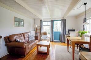 A living room with a brown leather sofa, a wooden coffee table, a dining table with chairs, a lamp, and a window with curtains. - A cozy stay near the beach, nature, and tulip fields! (Callantsoog)