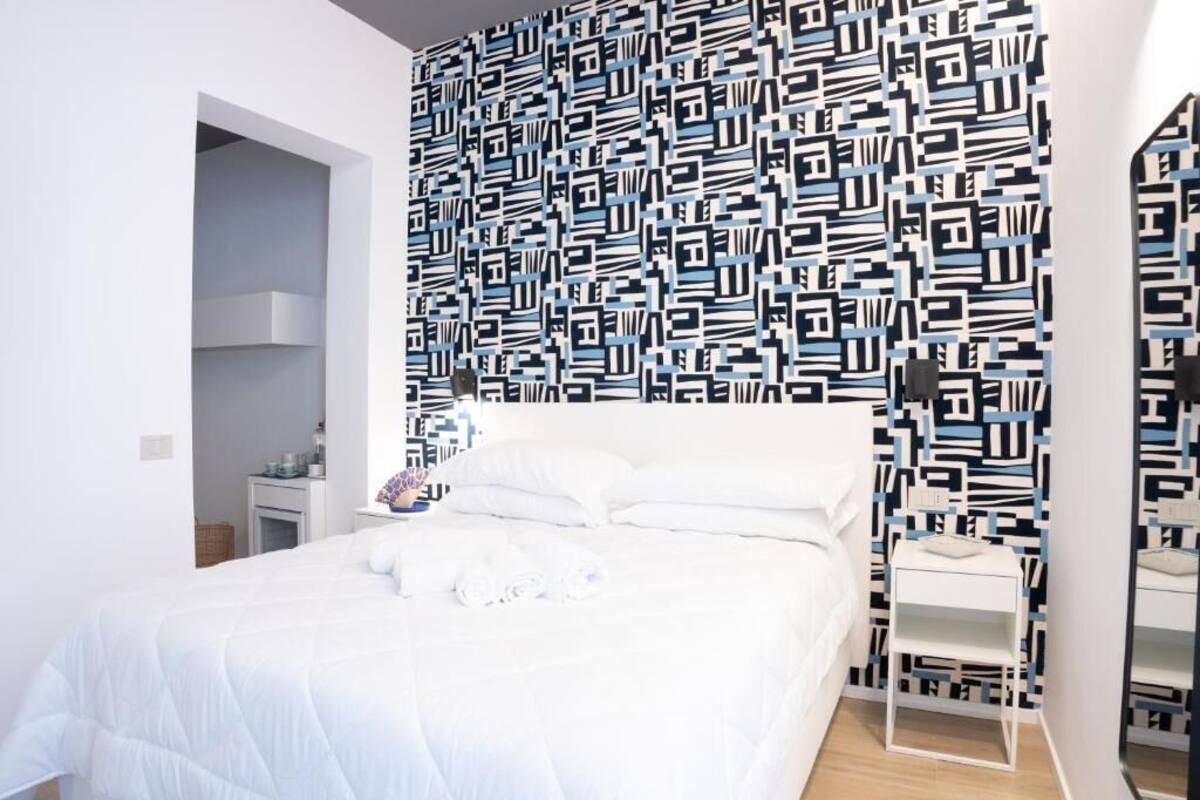 Un dormitorio con cama blanca, mesita de noche, espejo y pared con diseño.