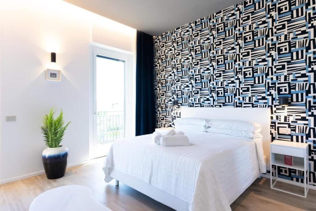 Un dormitorio moderno con pared estampada, una cama con ropa de cama blanca, una mesita de noche y una planta en maceta.
