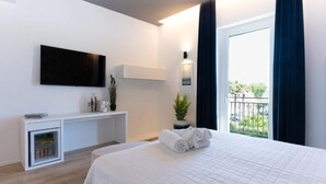 A hotel room with a bed, a TV mounted on the wall, a small desk with a plant, and a balcony with curtains.