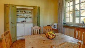 Uma sala de jantar com uma mesa de madeira, cadeiras, uma fruteira, uma cozinha com armários abertos e uma janela com cortinas.