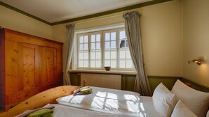 Quarto com guarda-roupa de madeira, cama com roupa de cama branca, janela com cortinas e radiador.