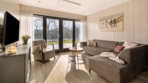 Ein modernes Wohnzimmer mit Sofa, Sesseln und Couchtisch, einem Flachbildfernseher und großen Fenstern mit Blick ins Freie.