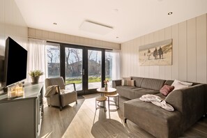 A modern living room with a sofa, armchair, and coffee table, a flat-screen TV, and large windows with a view of the outdoors. - Luxury & Relaxation at the Zee & Zand Family House<br> <br>Welcome to the Zee & Zand Family House—your brand-new vacation home, where luxury, comfort, and flexibility come together perfectly.<br>The accommodation consists of two beautiful 4-person— (Julianadorp)