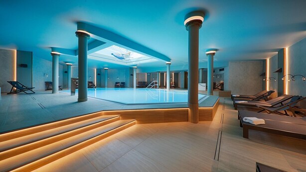 Un vaste espace intérieur abritant une piscine, avec une piscine centrale, de nombreux fauteuils de relaxation et un design architectural moderne agrémenté de colonnes et d’une verrière.