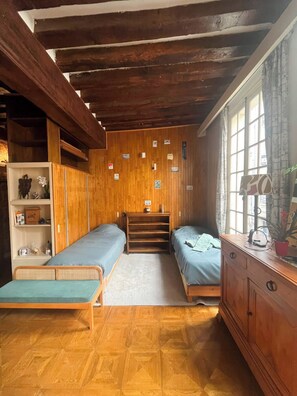 Ein Zimmer mit zwei Einzelbetten, Holzwänden, einem Fenster mit Vorhängen und einem Regal mit diversen Gegenständen.
